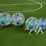 A group of students wearing inflatable bubble suits are playing a lively game of 5-a-side soccer on a green turf field. The participants, encased in transparent, spherical bubbles, bump into each other while energetically chasing the soccer ball. An official in a yellow jersey stands in the background. © Powerleague