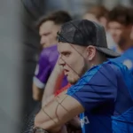 A man wearing a blue sports jersey and a black cap is leaning against a fence, watching a bubble football match intently. Several people are in the background, some of them wearing purple jerseys. The scene appears to be outdoors and sunny. © Powerleague