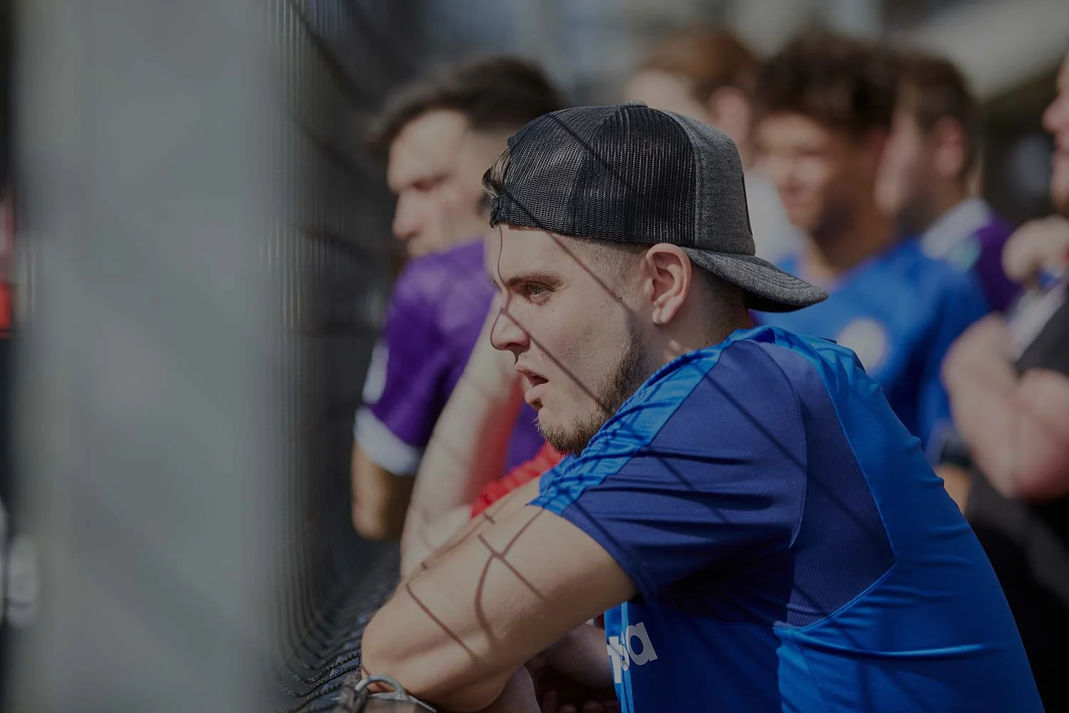 A man wearing a blue sports jersey and a black cap is leaning against a fence, watching a bubble football match intently. Several people are in the background, some of them wearing purple jerseys. The scene appears to be outdoors and sunny. © Powerleague