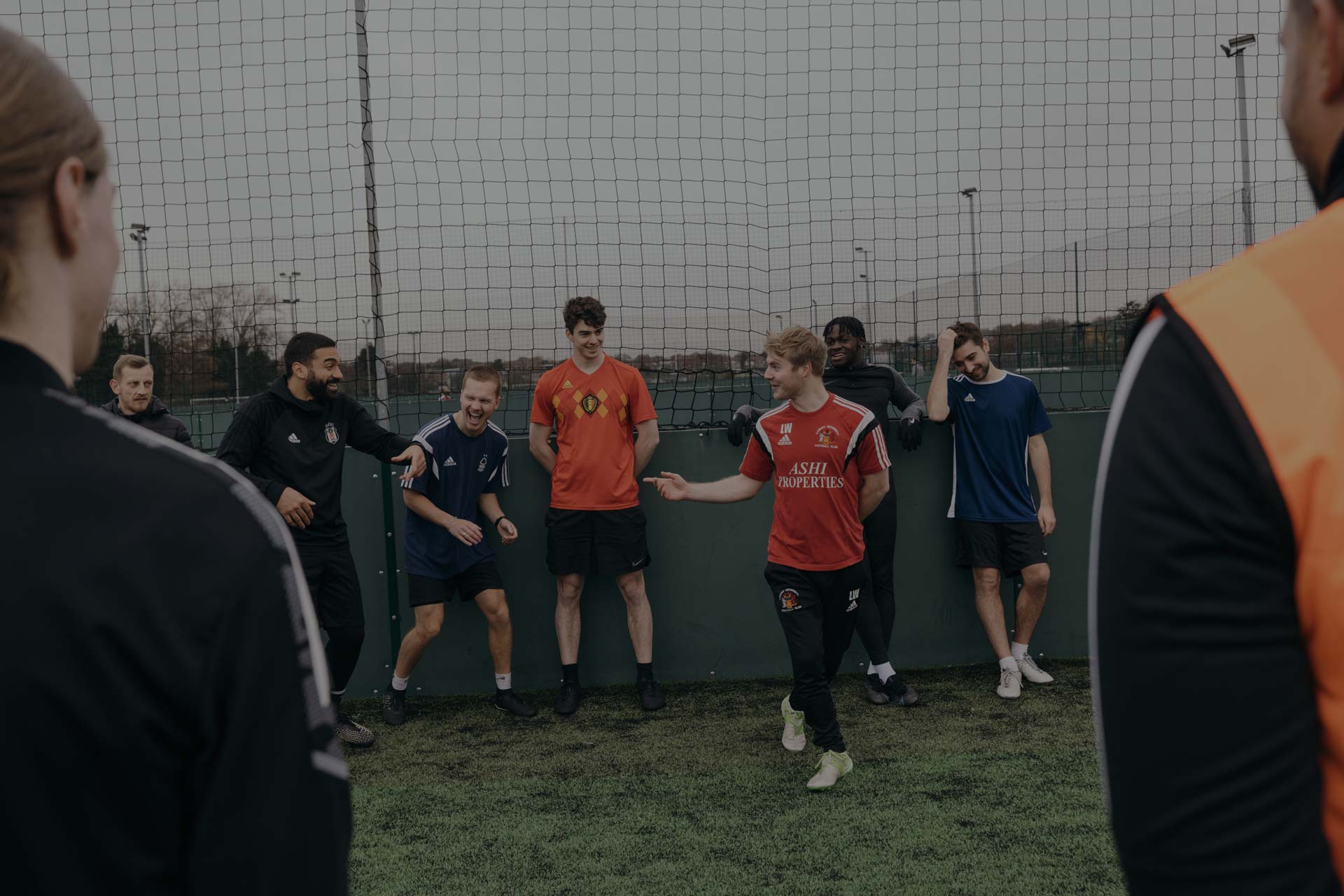 A group of students in athletic clothing are standing and chatting by a netted fence on a grassy field. One man in a red sports jersey is animatedly speaking to the 5-a-side team, while others smile and look on. Some individuals wear sports jackets, and the scene appears casual and friendly. © Powerleague