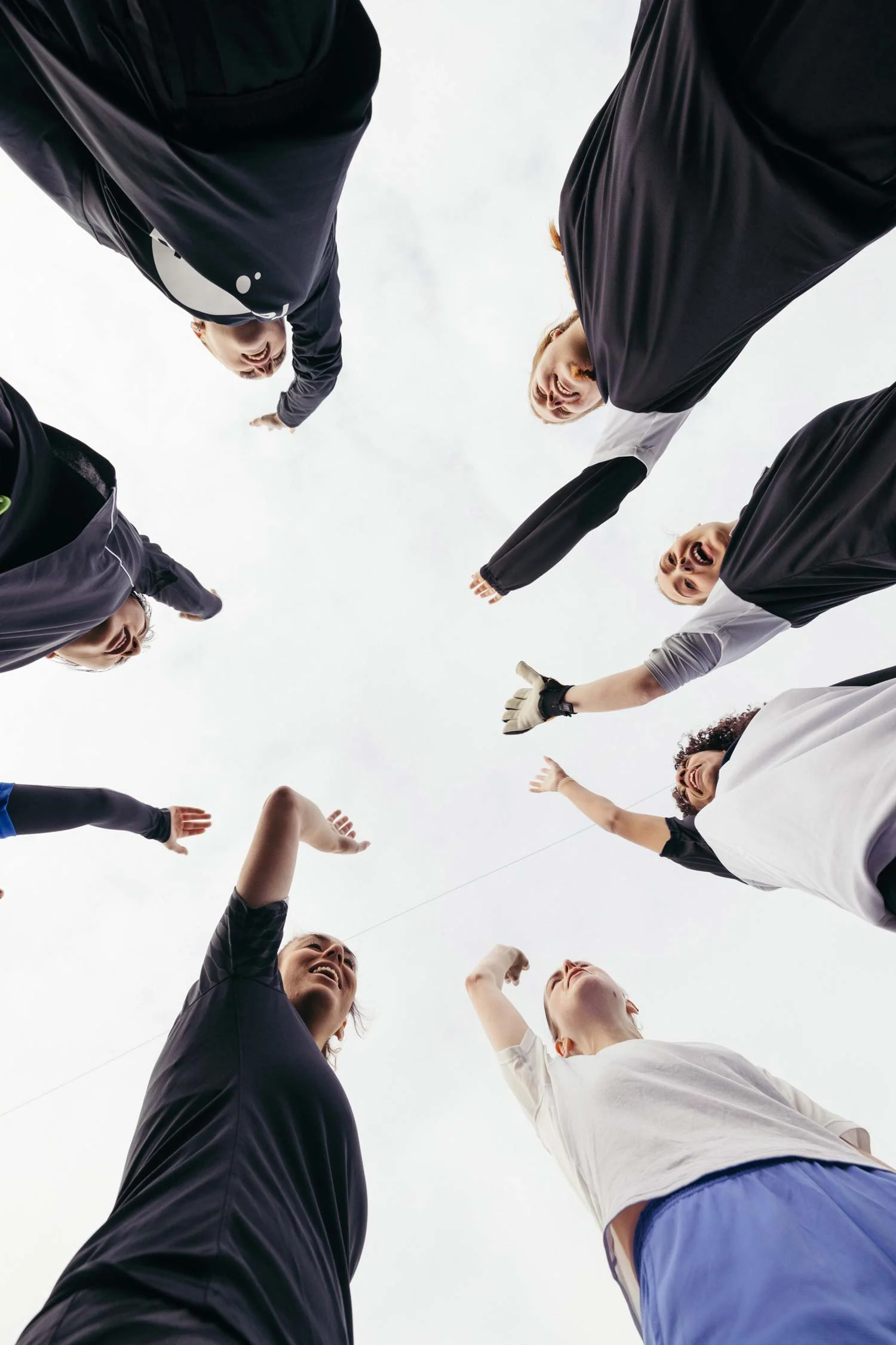 A group of people dressed in sportswear, including a women's football team, stand in a circle with their arms raised, viewed from below against a cloudy sky. The image captures a sense of unity and team spirit. © Powerleague