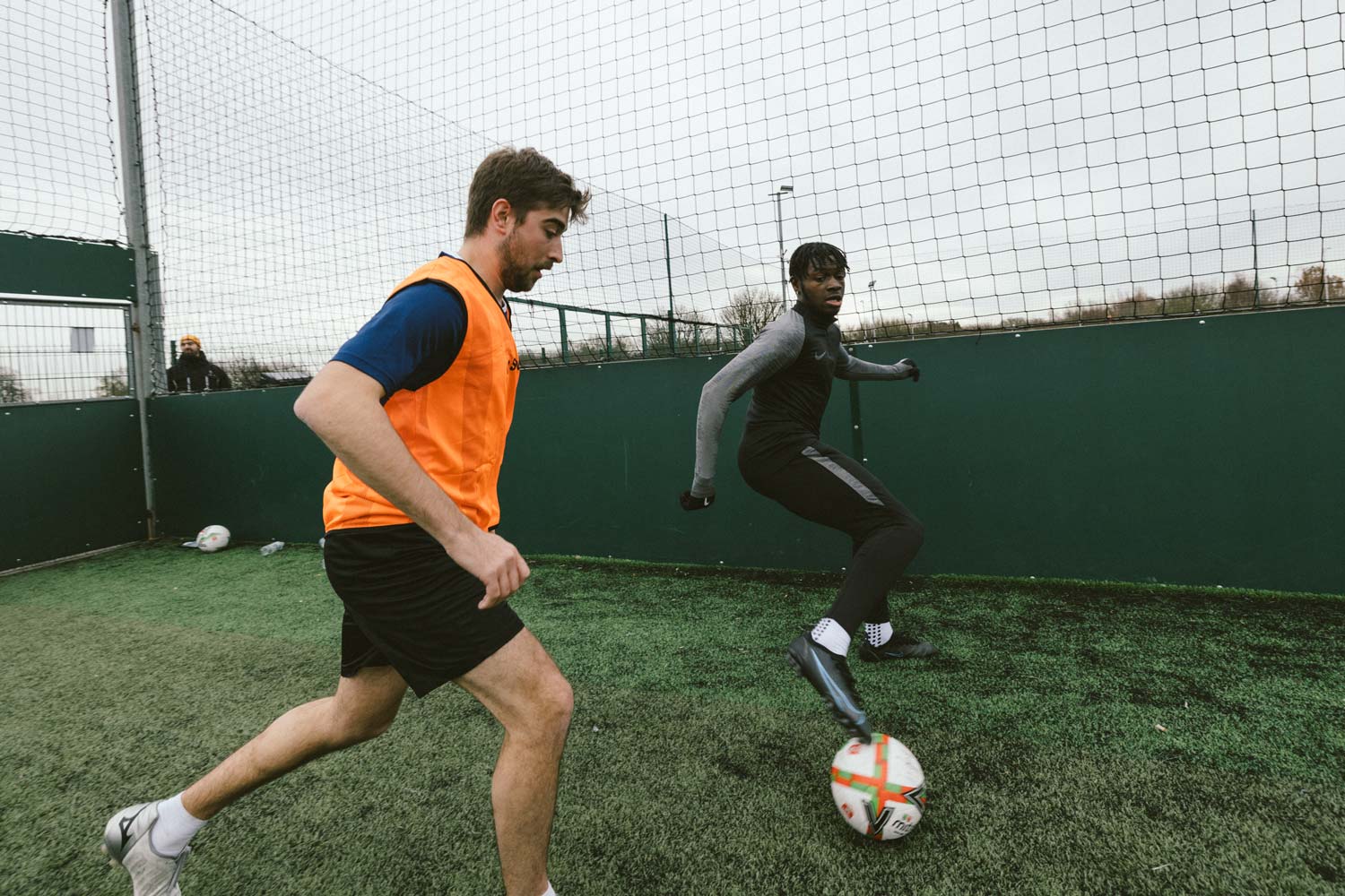 Two people are playing soccer on an outdoor field surrounded by a high fence. The player on the left, wearing an orange vest, is chasing the player on the right, who is wearing a black jacket and has a prosthetic leg. Both students are focused on the ball in front of them during this intense 5-a-side match. © Powerleague