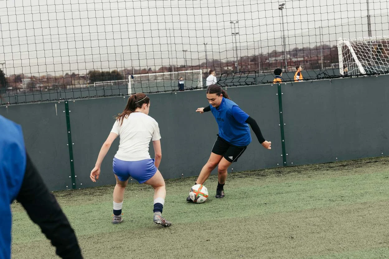 Two people playing soccer on an outdoor field. One, in a white shirt and blue shorts, defends while the other, in a blue shirt and black shorts, controls the ball. A goalkeeper in an orange vest stands in goal, backgrounded by a net and a cloudy sky.