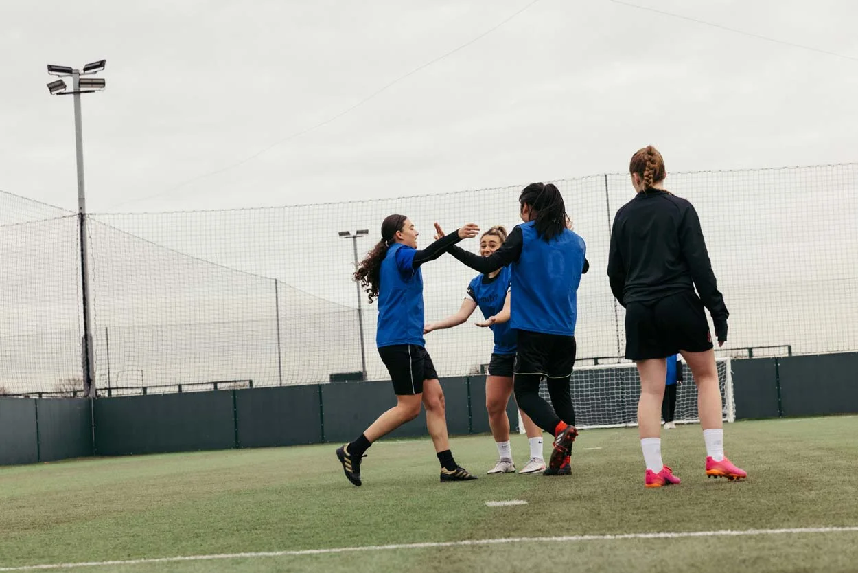 Four female soccer players are on a field celebrating. Three of the players wearing blue jerseys are high-fiving each other, while the fourth player in a black jersey stands nearby. The field is fenced and has tall lights in the background.
