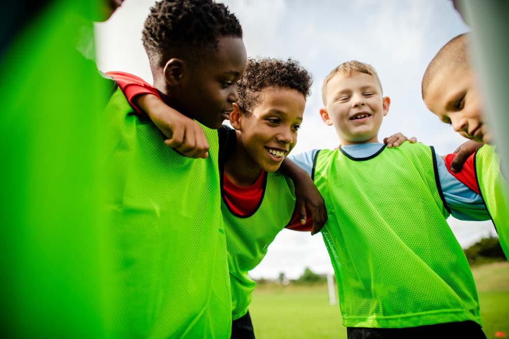 A group of young boys wearing bright green soccer jerseys huddle together with their arms around each other, smiling and laughing on a grassy field under a clear blue sky, reminiscent of the camaraderie found at football holiday camps. © Powerleague