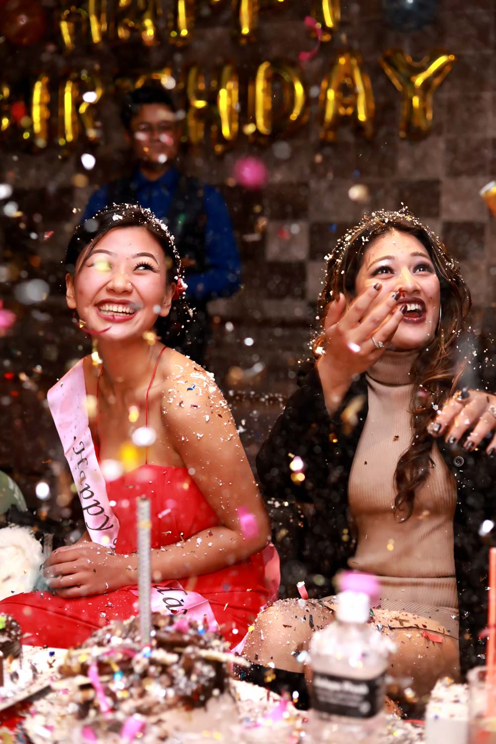Two smiling women celebrate a birthday amid a shower of confetti. One wears a pink dress and sash reading 