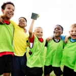 A group of six young boys, wearing green sports vests over their athletic clothes, stand together on a grassy field. They are smiling and looking happy, with one boy holding a small trophy. They have their arms around each other's shoulders in a celebratory pose. © Powerleague