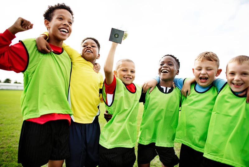 A group of six young boys, wearing green sports vests over their athletic clothes, stand together on a grassy field. They are smiling and looking happy, with one boy holding a small trophy. They have their arms around each other's shoulders in a celebratory pose. © Powerleague