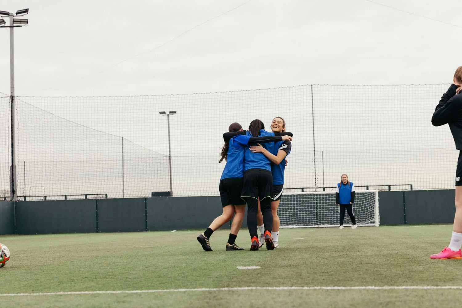 A group of three women's football players dressed in blue and black uniforms joyfully embrace on an outdoor soccer field. Another player, also in blue and black, watches them from near the goal. A coach or player stands in the background facing away. © Powerleague