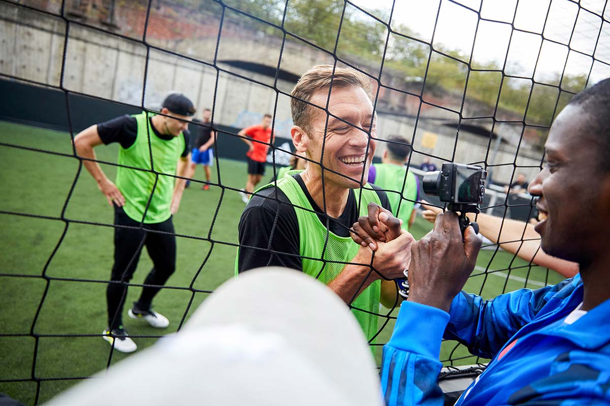 A man in a green sports jersey smiles and shakes hands with another person through a soccer net, highlighting the camaraderie of team building days. Other players in green and black jerseys are visible in the background on the field, as a camera captures the interaction. © Powerleague