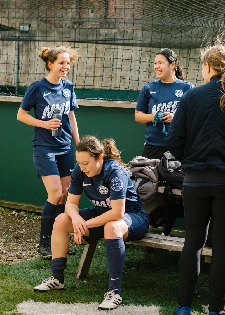 Four women in bubble football uniforms are gathered by the side of a field, taking a break. Two are standing and laughing, one is sitting on a bench looking down, and another has her back to the camera. They appear to be in a relaxed and cheerful mood. © Powerleague