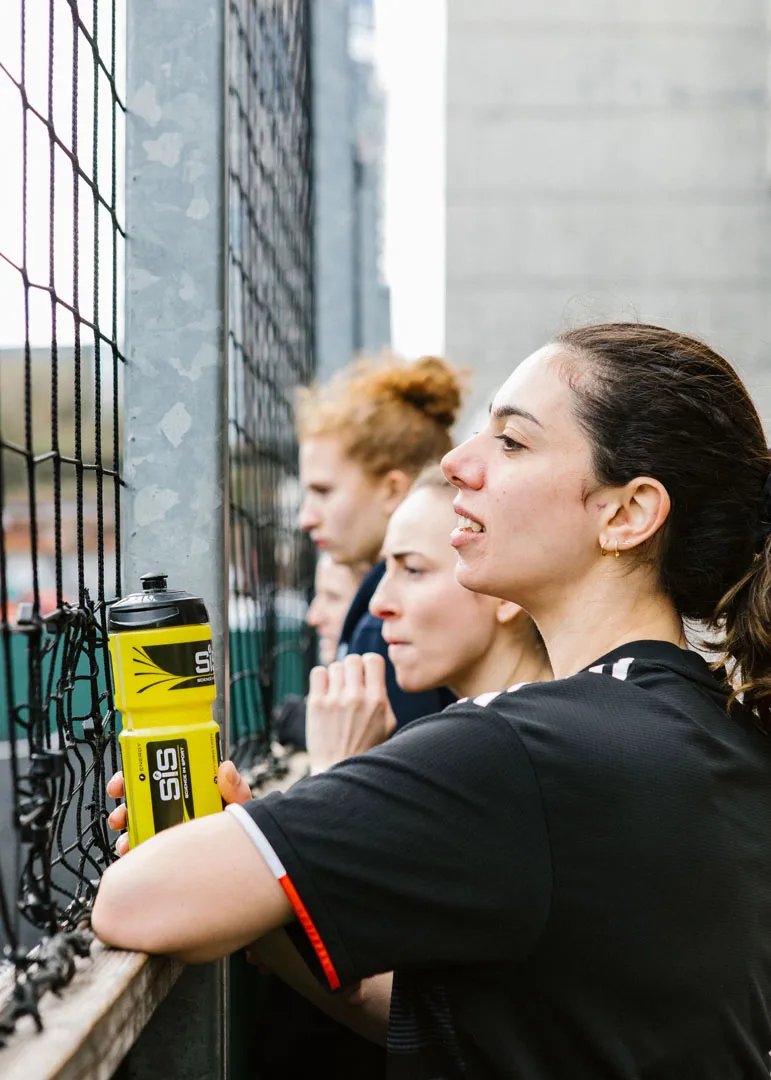 Three individuals are standing near a wire fence, looking in the same direction. Only one person's face is fully visible; she is holding a yellow water bottle. The others' faces are partially obscured. They appear to be watching something intently, perhaps a bubble football match in progress. © Powerleague