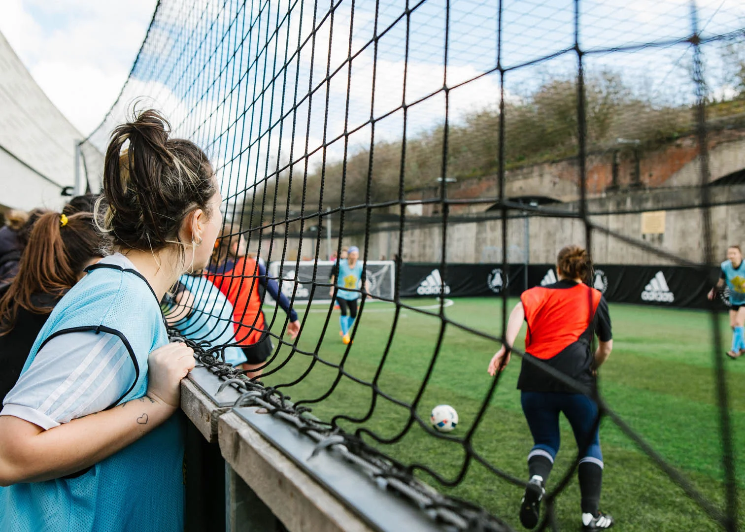 A group of people play women's football on an outdoor field surrounded by tall netting. One person wearing a light blue bib leans on the fence, watching the game. Other players are wearing red and blue bibs, actively engaged in the match. © Powerleague