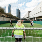 A soccer match takes place in an urban sports facility with skyscrapers in the background. A female goalkeeper in a neon green vest and black shorts is seen from behind, watching the bubble football action as players compete for the ball in front of the goal net. © Powerleague