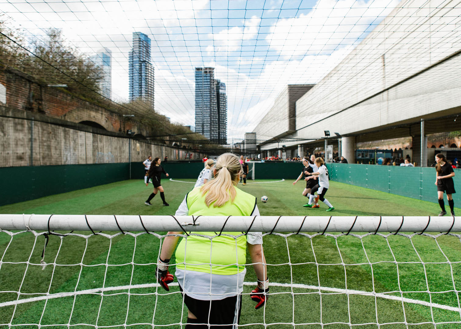 A soccer match takes place in an urban sports facility with skyscrapers in the background. A female goalkeeper in a neon green vest and black shorts is seen from behind, watching the bubble football action as players compete for the ball in front of the goal net. © Powerleague