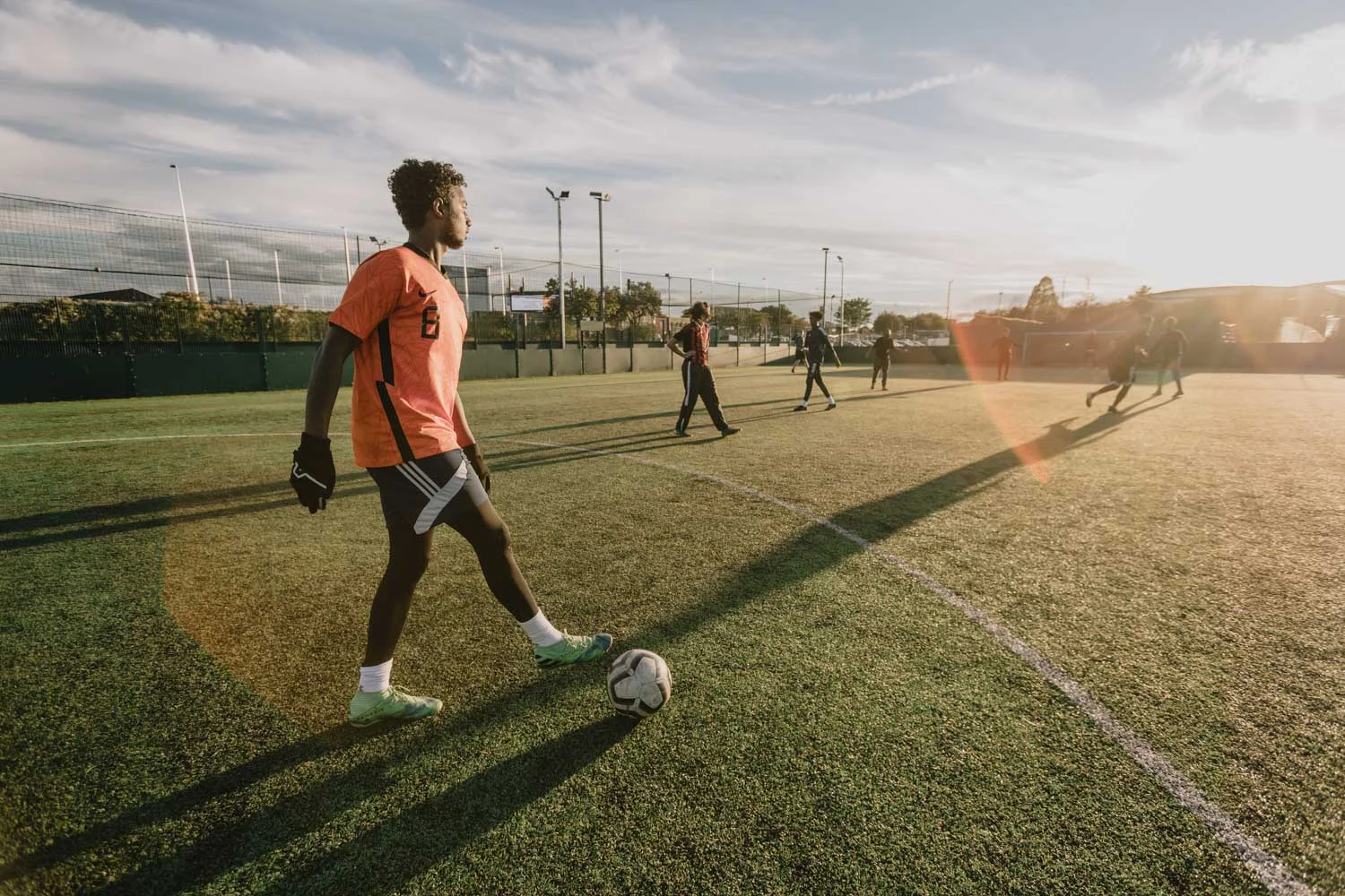 A person wearing an orange shirt and black shorts stands on a soccer field with a ball at their feet, preparing to play. The sun is low in the sky, casting long shadows, with several other players from local football leagues visible in the background. © Powerleague