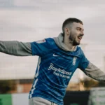 A man with short hair and a beard, wearing a blue Birmingham City football jersey over a grey hoodie, is enthusiastically celebrating with his arms outstretched. He appears to be on a field, possibly part of a student 5-a-side game, with a net and blurred background visible behind him. © Powerleague