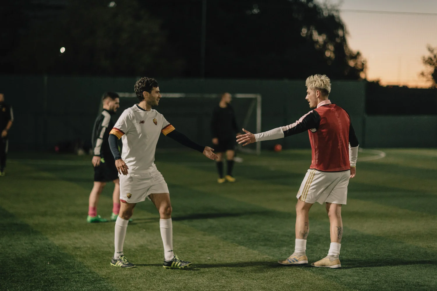 Two football players, one in a white uniform with a captain's armband and the other in a red training bib over a white shirt, meet for a handshake on an outdoor field. Other players and a goalpost are visible in the background during what appears to be early evening, typical of 5-a-side football. © Powerleague