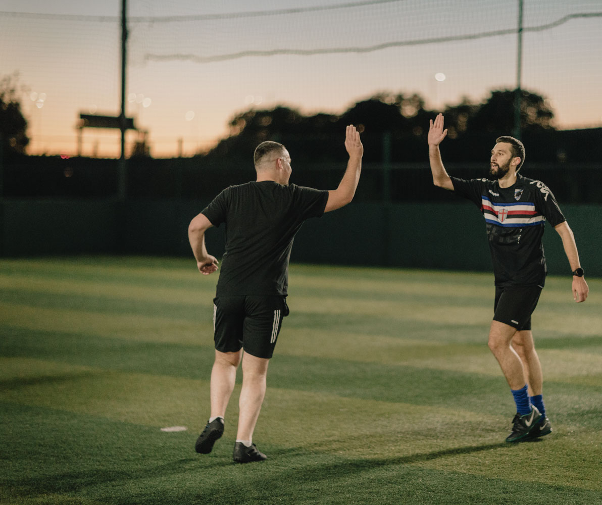 Two men wearing sports attire high-five on a soccer field during sunset. One wears a black shirt and shorts, while the other, resembling veterans of the game, dons a black sports jersey with white, blue, and red stripes, and black shorts. The field is enclosed with netting, and trees are visible in the background. © Powerleague