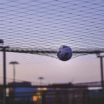 A soccer ball caught in mid-air within a net, against a backdrop of a cityscape at dusk with soft purple and orange hues in the sky. A few blurry structures and lights are visible in the background. © Powerleague