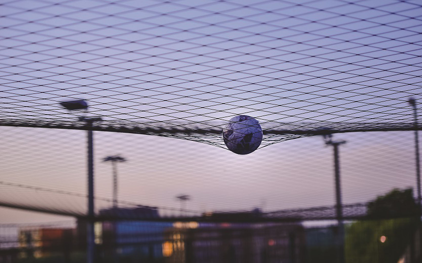 A soccer ball caught in mid-air within a net, against a backdrop of a cityscape at dusk with soft purple and orange hues in the sky. A few blurry structures and lights are visible in the background. © Powerleague