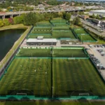 Aerial view of a sports facility with multiple fenced soccer fields, including areas for 5-a-side football. Players are seen on the fields, and a central building is surrounded by parking lots. Nearby, a river runs parallel to a highway with overpasses and vehicles. Industrial buildings are visible in the background. © Powerleague