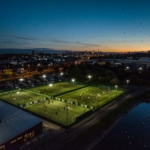 Aerial view of a sports complex with multiple lit-up soccer fields at dusk. Players from local football leagues are seen on the fields. The cityscape glows in the distance under the darkening sky, with birds flying overhead and a river adjacent to the complex. © Powerleague