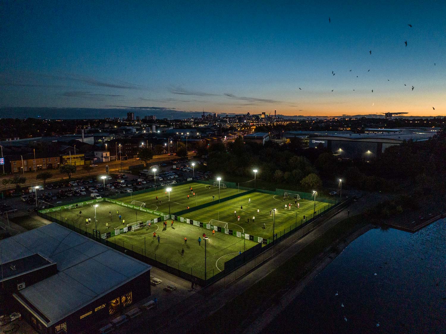 Aerial view of a sports complex with multiple lit-up soccer fields at dusk. Players from local football leagues are seen on the fields. The cityscape glows in the distance under the darkening sky, with birds flying overhead and a river adjacent to the complex. © Powerleague