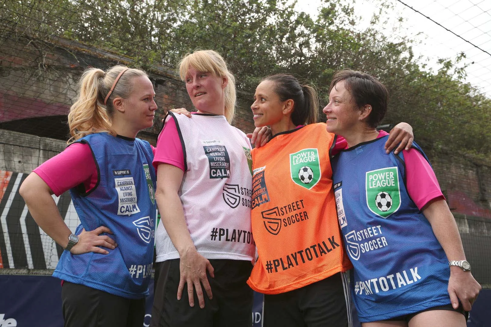 Four women in blue and orange Street Soccer vests stand close together, smiling and looking at each other. They are on an outdoor soccer field with trees and a wire fence in the background, participating in a #PlayToTalk women's football event. © Powerleague
