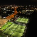 Aerial nighttime view of a sports complex with multiple illuminated soccer fields, resembling a bustling transfer market. Surrounding city streets and buildings are visible, with streetlights and vehicle lights creating a network of glowing lines. © Powerleague