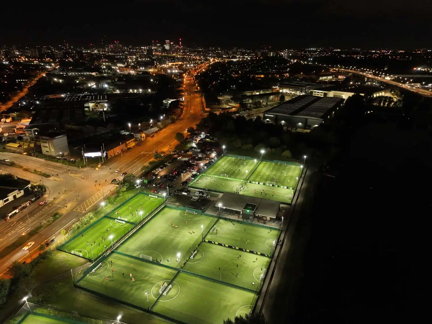 Aerial nighttime view of a sports complex with multiple illuminated soccer fields, resembling a bustling transfer market. Surrounding city streets and buildings are visible, with streetlights and vehicle lights creating a network of glowing lines. © Powerleague