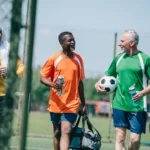 Four men in sportswear are walking on a soccer field, perhaps enjoying a lively game of walking football. Three are holding water bottles and one has a soccer ball. They are smiling and appear to be engaged in conversation. A fence and goalpost are visible in the background. © Powerleague