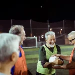 A group of older men is enjoying an evening game of walking football on a well-lit field. They are in high spirits, sharing a laugh, and one man in a green bib is holding a soccer ball. The background features a goalpost and a netted fence. © Powerleague