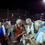 A group of five elderly men wearing sports attire and bibs are sitting on a football field at night, smiling and chatting with one another. Having just finished a walking football game, they have towels around their necks. The field is surrounded by net fencing. © Powerleague