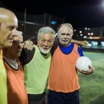 A group of elderly men, wearing brightly colored sports jerseys, stand together on a soccer field at night. With arms around each other, they smile and hold a soccer ball, illuminated by the stadium lights in the background, ready for their game of walking football. © Powerleague