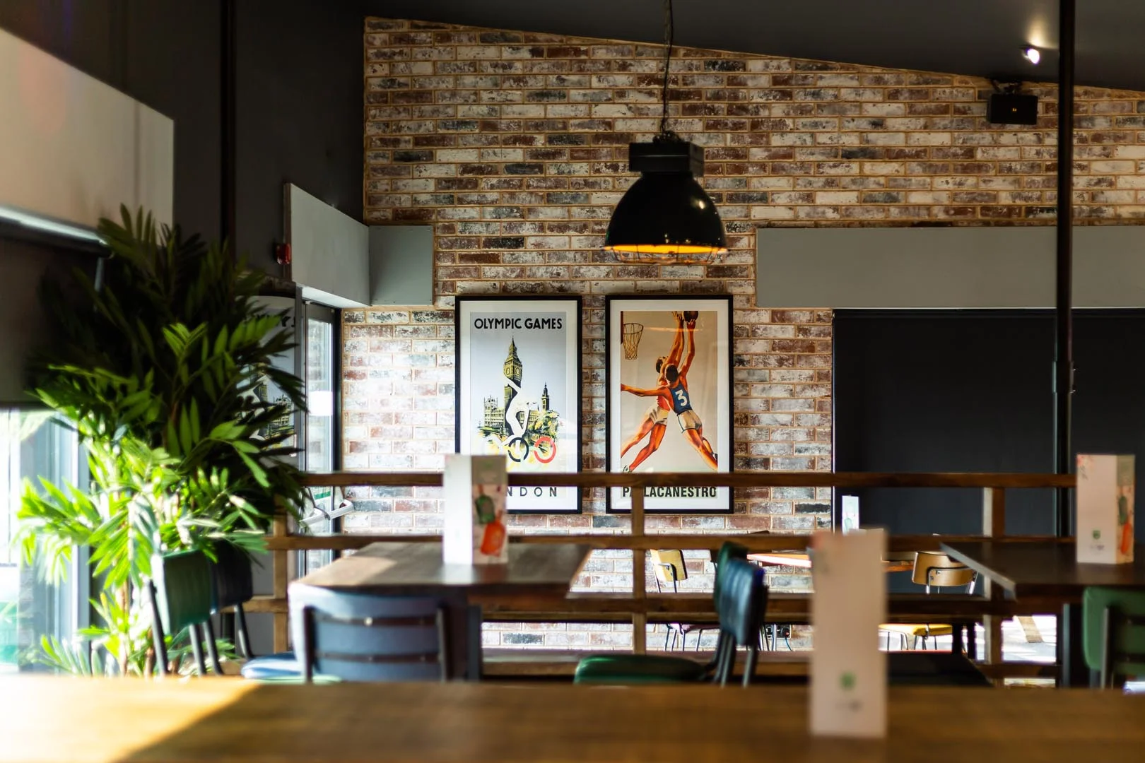 A modern cafe with brick walls, framed Olympic Games posters, and large windows. There are plants, pendant lighting, and wooden tables with menus. Various types of seating are visible, including high stools and chairs. The bright and inviting atmosphere makes it a perfect spot for bubble football enthusiasts to gather. © Powerleague