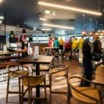 A lively bar with modern, industrial décor, featuring wooden tables and chairs, hanging lights, and a bar counter in the background. People are standing and socializing near the bar while discussing their recent bubble football match. The space is well-lit and features exposed brick and large windows. © Powerleague