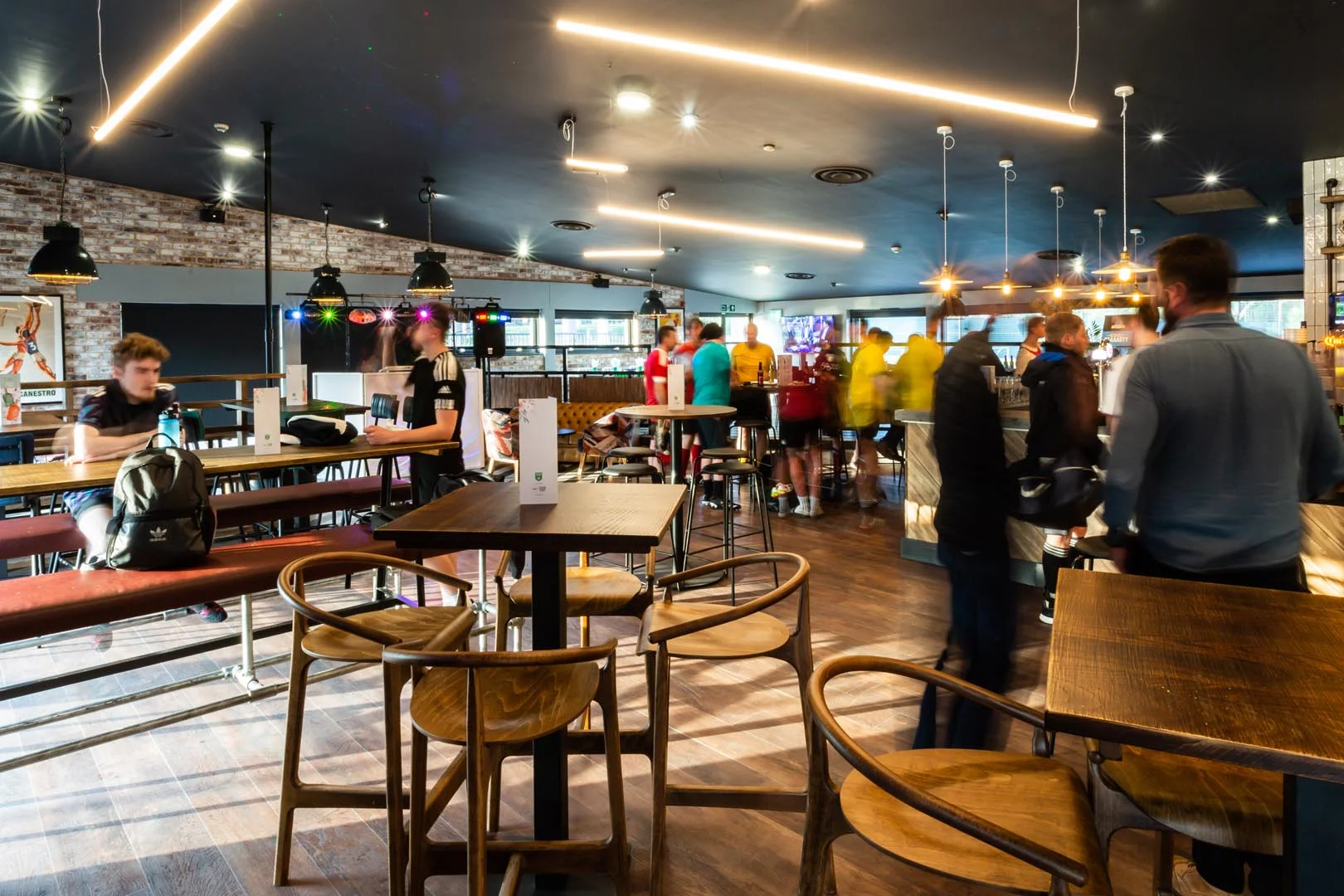 A lively bar with modern, industrial décor, featuring wooden tables and chairs, hanging lights, and a bar counter in the background. People are standing and socializing near the bar while discussing their recent bubble football match. The space is well-lit and features exposed brick and large windows. © Powerleague