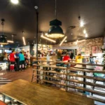 A lively café with customers at the bar and tables, some wearing sports jerseys from their bubble football games. Modern decor includes pendant lights, wooden furniture, a brick accent wall, and a plant. Large windows let in natural light, highlighting the bustling, friendly atmosphere. © Powerleague