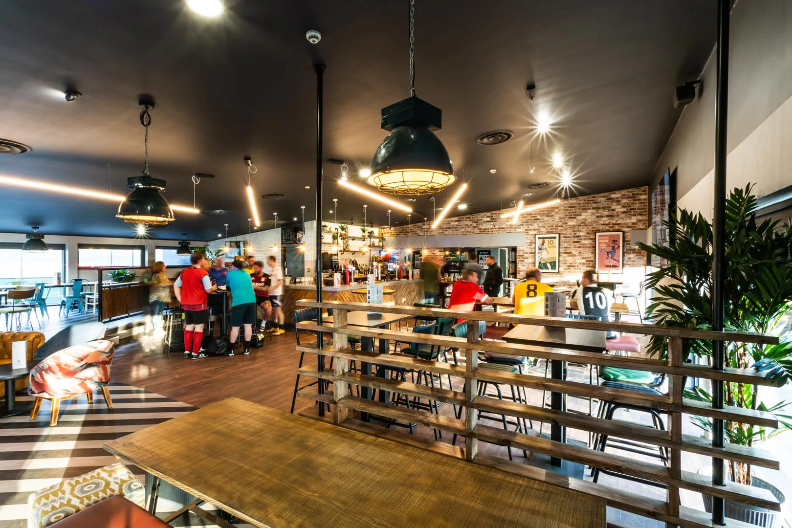 A lively café with customers at the bar and tables, some wearing sports jerseys from their bubble football games. Modern decor includes pendant lights, wooden furniture, a brick accent wall, and a plant. Large windows let in natural light, highlighting the bustling, friendly atmosphere. © Powerleague