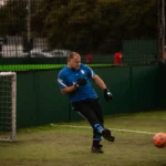A soccer player wearing a blue jersey and black pants kicks a ball on an indoor veterans football field. The goalpost is visible to the left, while an orange ball is in mid-air. The background shows a green fence and blurred figures. © Powerleague