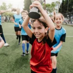 A group of children in soccer uniforms are celebrating on a 5-a-side football field. A girl in a red jersey is at the center holding up a trophy with a big smile, while her teammates in blue jerseys cheer behind her. Everyone looks joyful and triumphant. © Powerleague