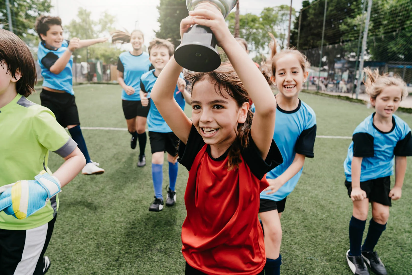 A group of children in soccer uniforms are celebrating on a 5-a-side football field. A girl in a red jersey is at the center holding up a trophy with a big smile, while her teammates in blue jerseys cheer behind her. Everyone looks joyful and triumphant. © Powerleague