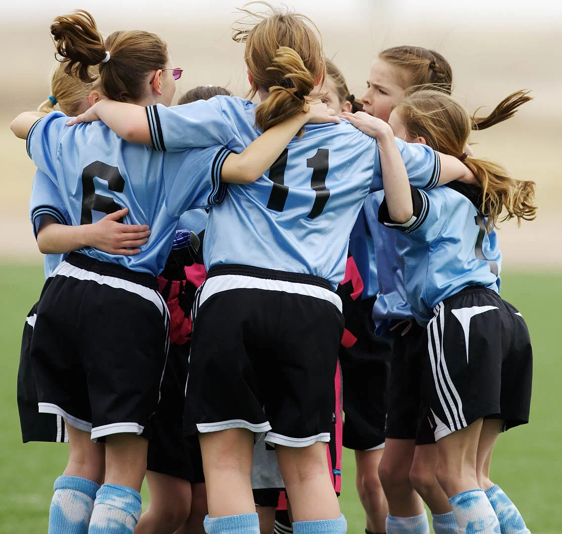 A group of young female soccer players in blue jerseys huddle together on a soccer field. They have their arms around each other, showing team spirit and camaraderie during their junior football training. The players have different jersey numbers visible, including 6 and 11. © Powerleague