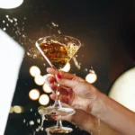 A hand with red painted nails holds a martini glass filled with a golden liquid, possibly a cocktail. The liquid is splashing out, creating droplets against a blurred background with circular light spots, reminiscent of the energy and excitement of 5-a-side football, giving a lively and dynamic feel. © Powerleague