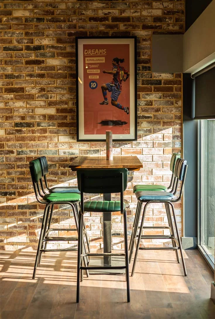 A small high-top table with four barstools, each featuring green cushions, is set against a brick wall with a framed art piece depicting a person in a bubble football jersey. Warm sunlight streams through large windows, casting light and shadows on the wooden floor. © Powerleague