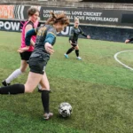A group of women play soccer on an artificial turf field. One player, at the center, is about to kick the ball while others are positioned around her in action. A banner in the background reads, 