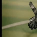 A close-up of a person's hand wearing black and white goalkeeper gloves on a green soccer field during a 5-a-side football match. The glove has a strap with the Adidas logo visible. The background is blurred, emphasizing the outstretched glove in the foreground. © Powerleague