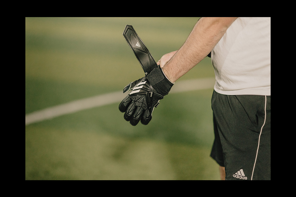 A person wearing a white shirt and black shorts is adjusting a black and white goalkeeper glove on their left hand. The blurred sports field in the background suggests a 5-a-side football environment. The person's face is not visible. © Powerleague