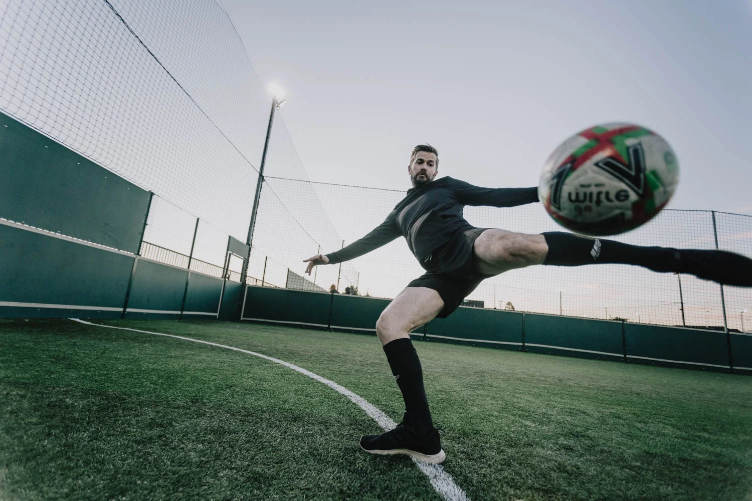 A man in athletic attire kicks a soccer ball mid-air on an outdoor 5-a-side football field with green synthetic turf. The sky is clear, and the field is surrounded by a tall netted fence. The angle of the shot is low, emphasizing the action and motion. © Powerleague